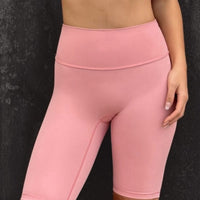 Woman wearing a whire sports bra and pink leggings against a dark textured wall.